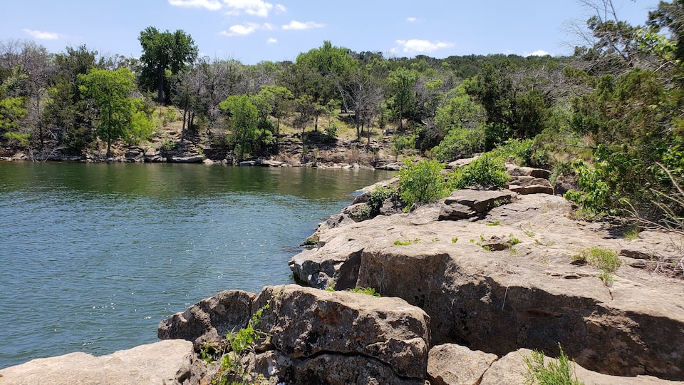 A rocky shoreline with a calm lake, surrounded by dense greenery and trees.