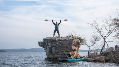 Eine Person steht auf einem Felsen, ein Paddel ist über dem Kopf, ein Kajak und ruhiges Gewässer sind zu sehen.