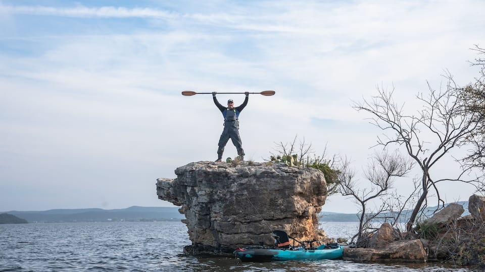 A person standing on a rock with a paddle over their head, a kayak, and a calm body of water.