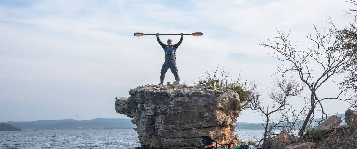 A person standing on a rock with a paddle over their head, a kayak, and a calm body of water.