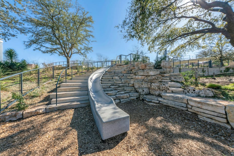 A curved stone staircase with a metal railing, surrounded by trees and a stone wall.