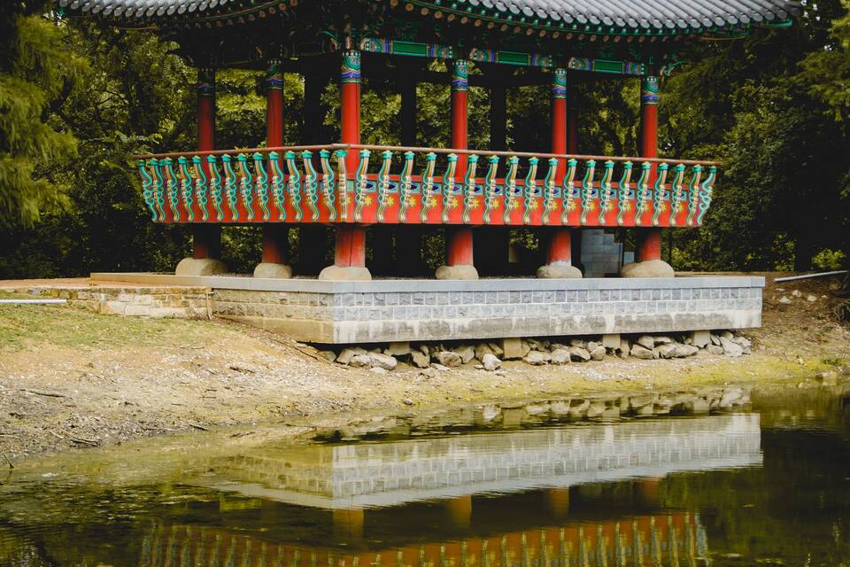 A traditional pavilion with a tiled roof, red pillars, and a stone base, reflecting in a pond surrounded by trees.