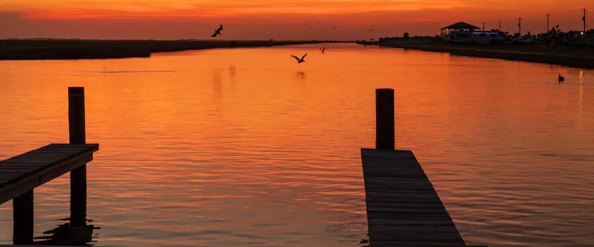 A breathtaking sunset casts a warm, orange glow over a calm river at Surfside Beach, Texas. The sky is painted with vibrant hues of orange and pink as the sun dips below the horizon.