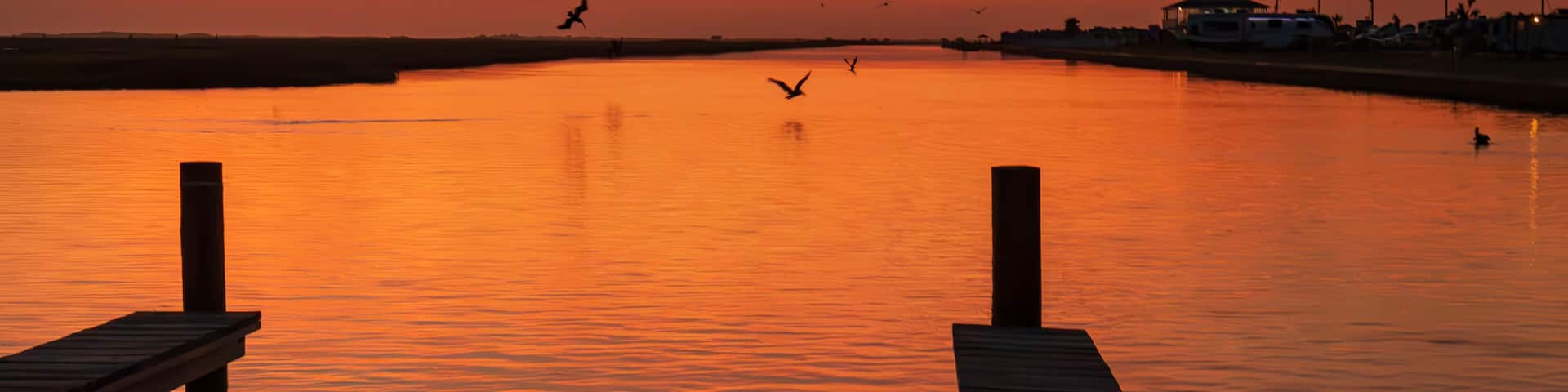 A breathtaking sunset casts a warm, orange glow over a calm river at Surfside Beach, Texas. The sky is painted with vibrant hues of orange and pink as the sun dips below the horizon.