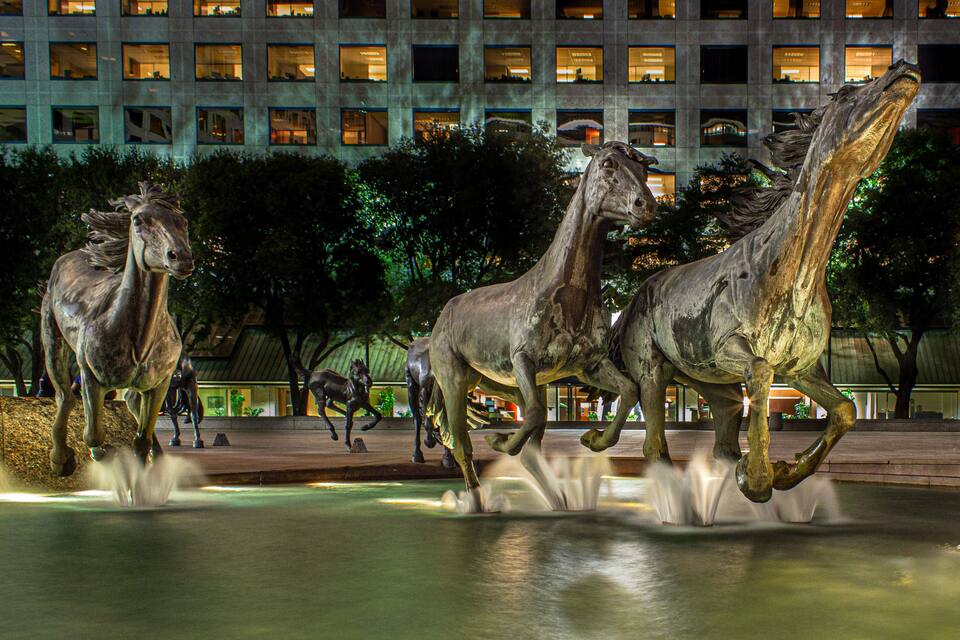 A nighttime sculpture of horses in motion, set against a backdrop of a modern building with illuminated windows.