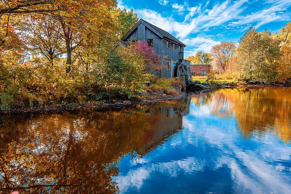 A wooden mill by a calm river, surrounded by autumn trees reflecting in the water.