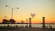 Un atardecer sobre un río con bancos de parque, farolas y torres decorativas.