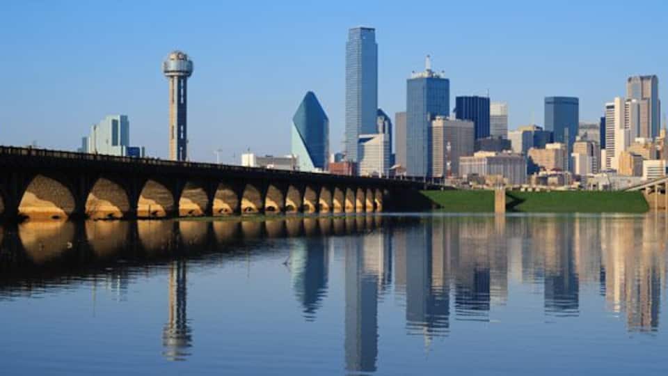 A city skyline with modern skyscrapers, a bridge over a calm river, and a prominent tower.