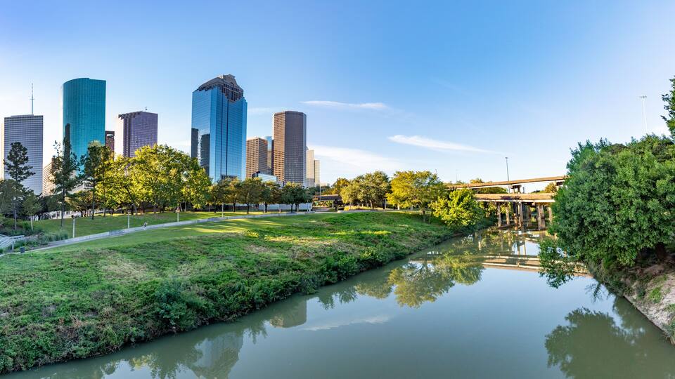 Um rio refletindo edifícios, um parque com árvores e uma ponte.