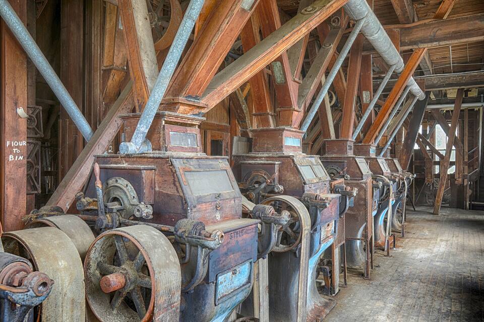 Een reeks oude, verroeste industriële machines met houten balken en metalen leidingen in een grote, zwak verlichte ruimte.