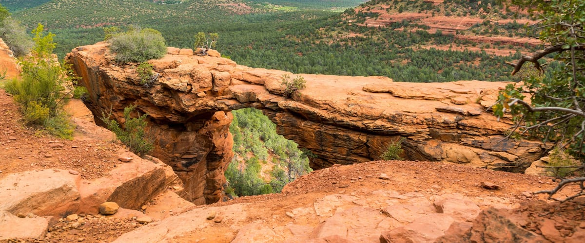 A rock arch formation in a desert landscape with surrounding hills and a cloudy sky.