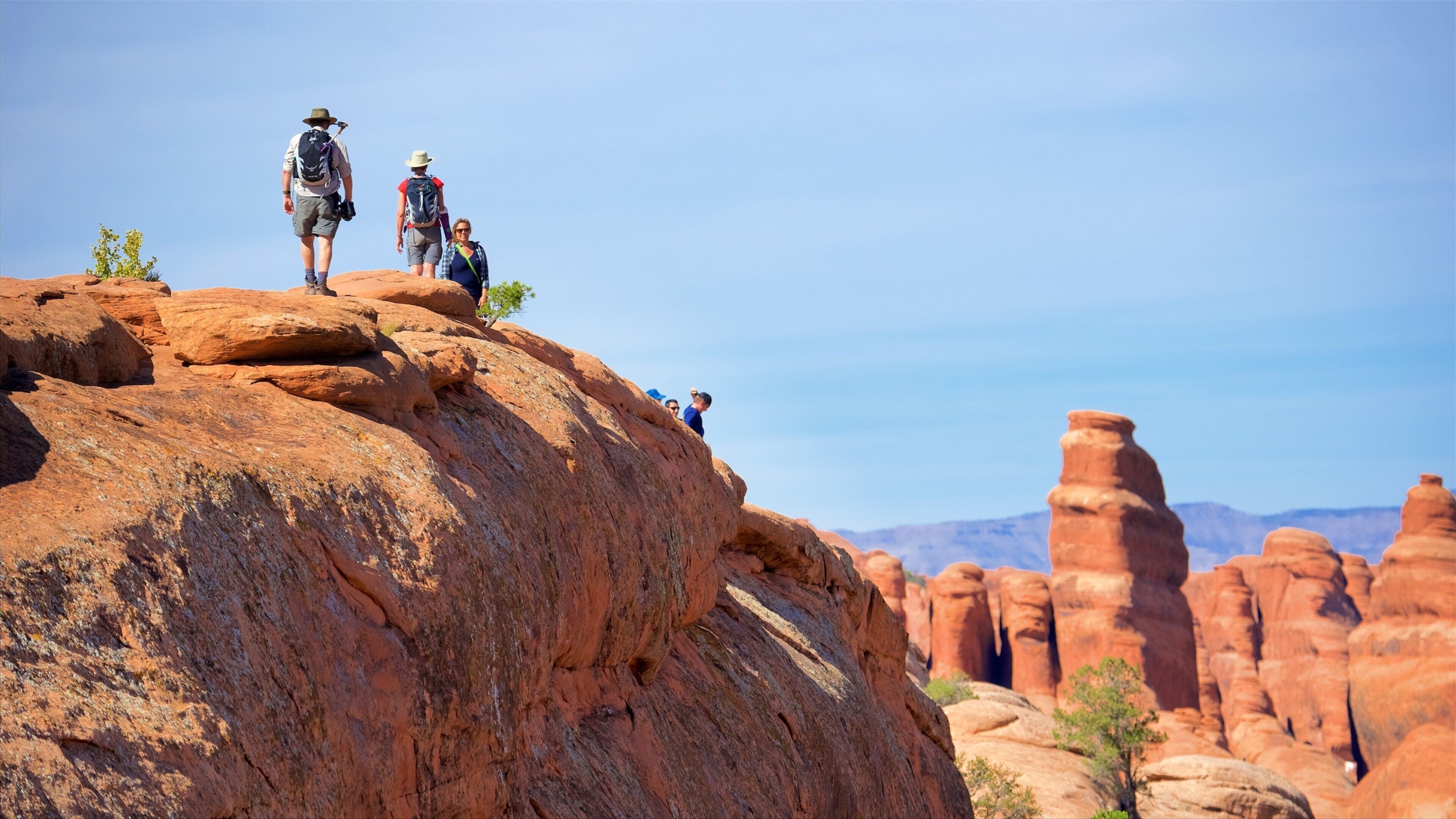 Hikers on a red rock formation with a clear sky and distant mountains.