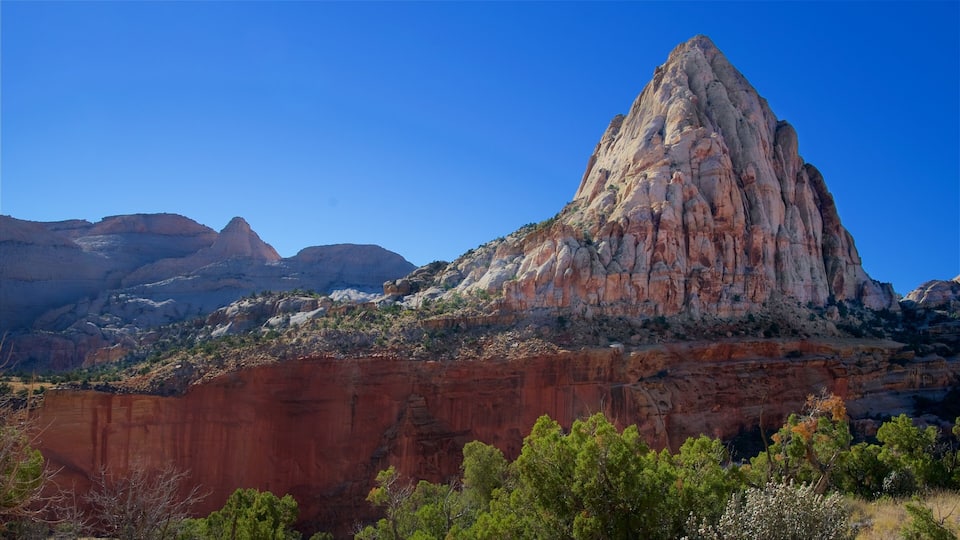 A prominent red rock formation with a peak, surrounded by green vegetation and other rocky mountains in the background.