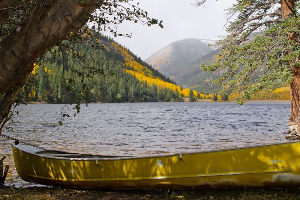 A yellow canoe tied to a tree by a calm lake with autumn-colored trees and mountains in the background.