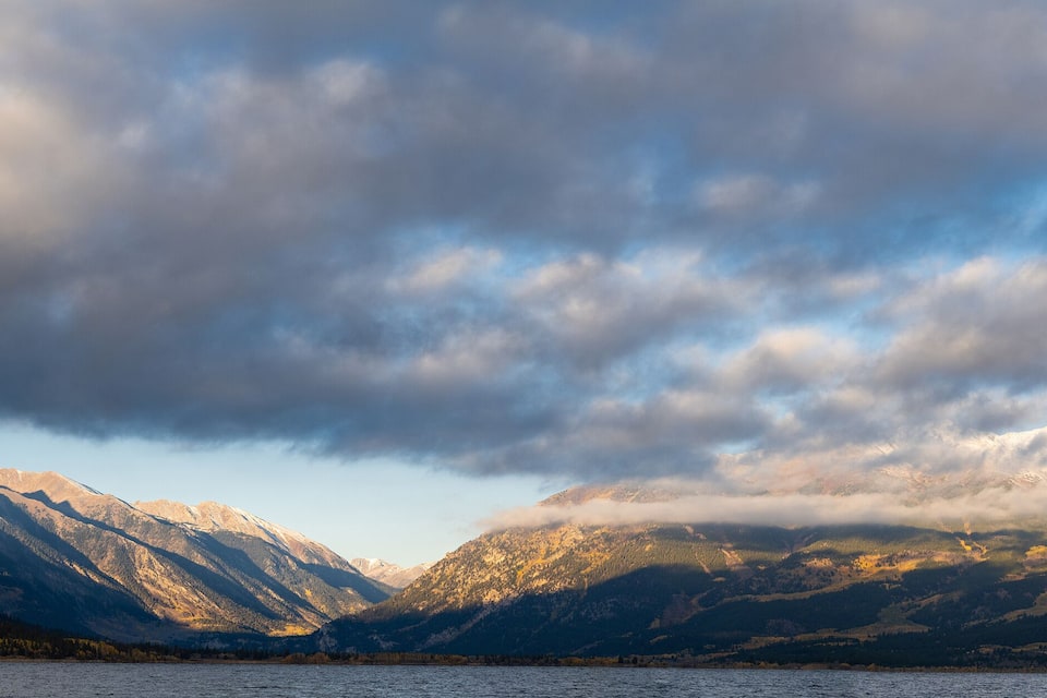 A mountain range with a lake in the foreground and a partly cloudy sky.