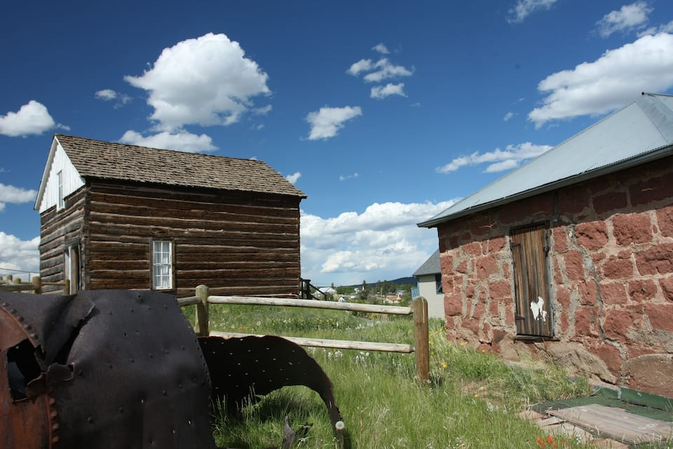 A rustic wooden cabin and a stone building with a metal door, surrounded by a wooden fence and a rusty metal object in a grassy field.