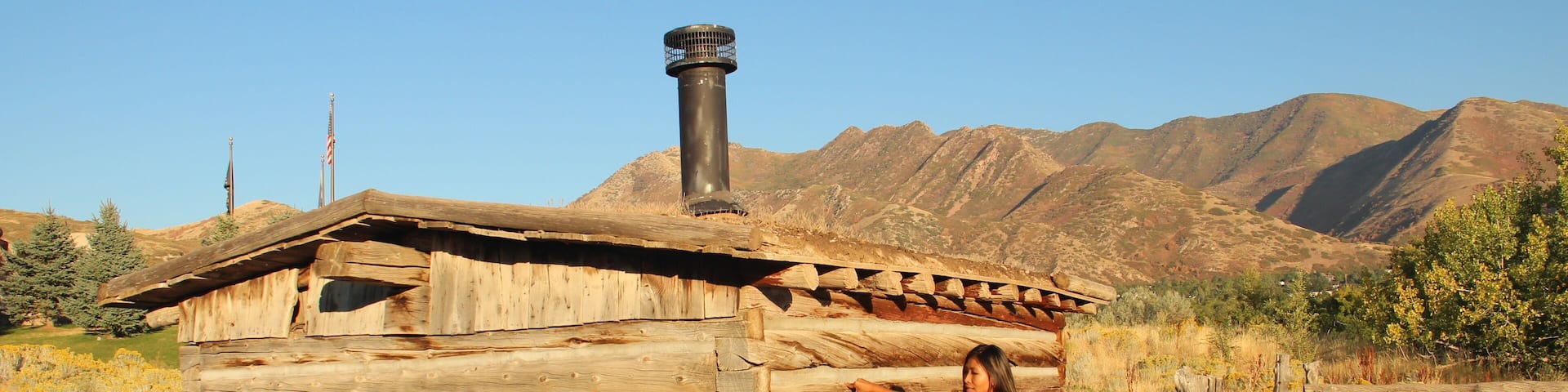 A wooden cabin with a chimney, surrounded by a fence and mountains in the background.