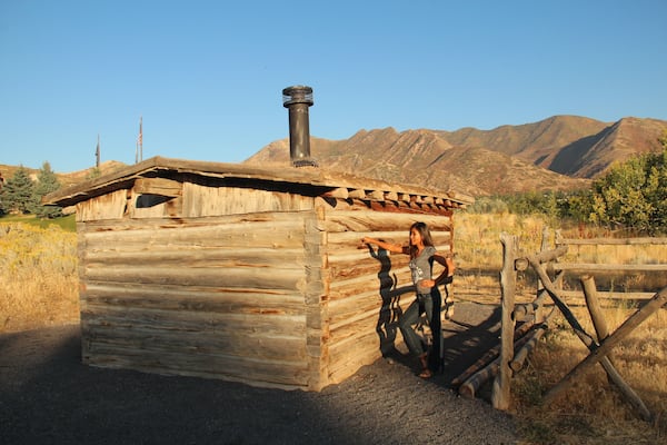 Une cabane en bois avec une cheminée, entourée d’une clôture, et des montagnes en arrière-plan.