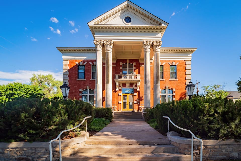 A red brick building with white columns and a blue door, surrounded by greenery.