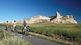 Trois cyclistes sur une piste asphaltée, avec un champ herbeux et une montagne rocheuse en arrière-plan.