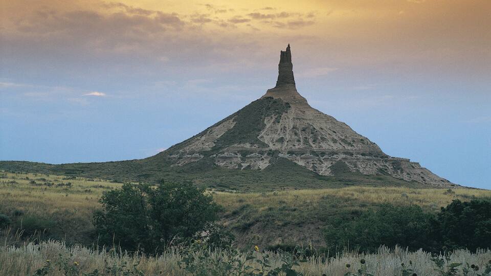 A prominent mountain peak with a distinct conical shape, surrounded by a grassy landscape with scattered trees and wildflowers.