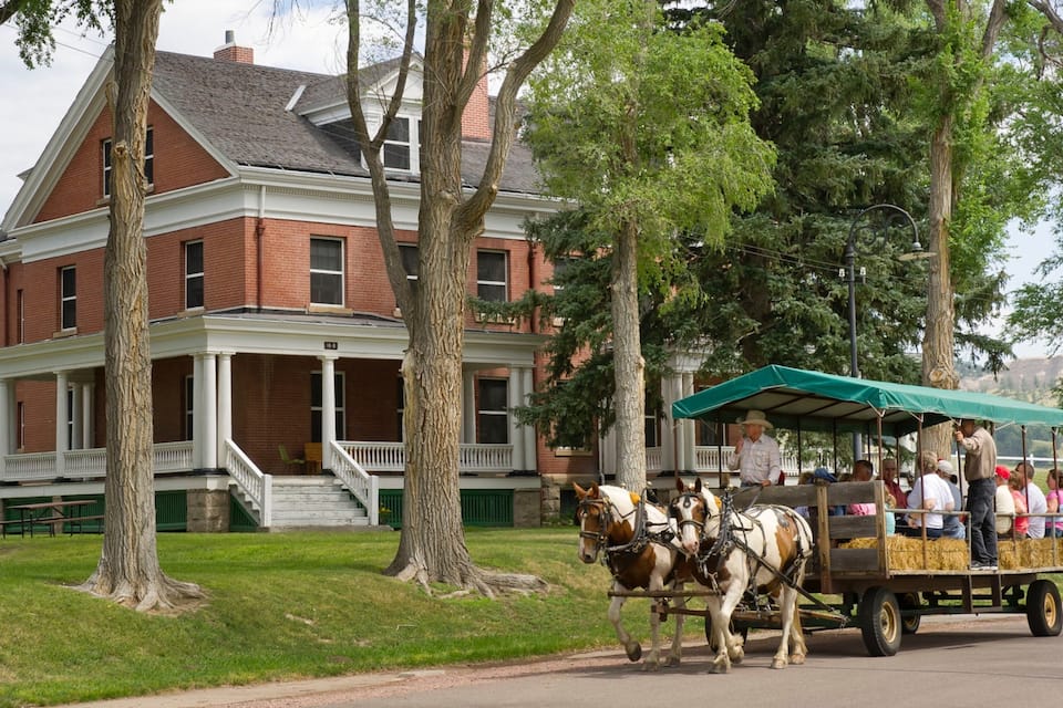 A horse-drawn carriage with hay bales, parked in front of a large brick building with a porch.