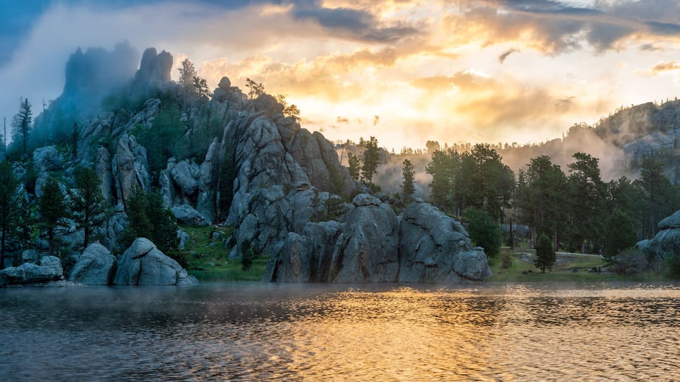 A mountainous landscape with a lake reflecting the sky, surrounded by dense forests and rocky outcrops.