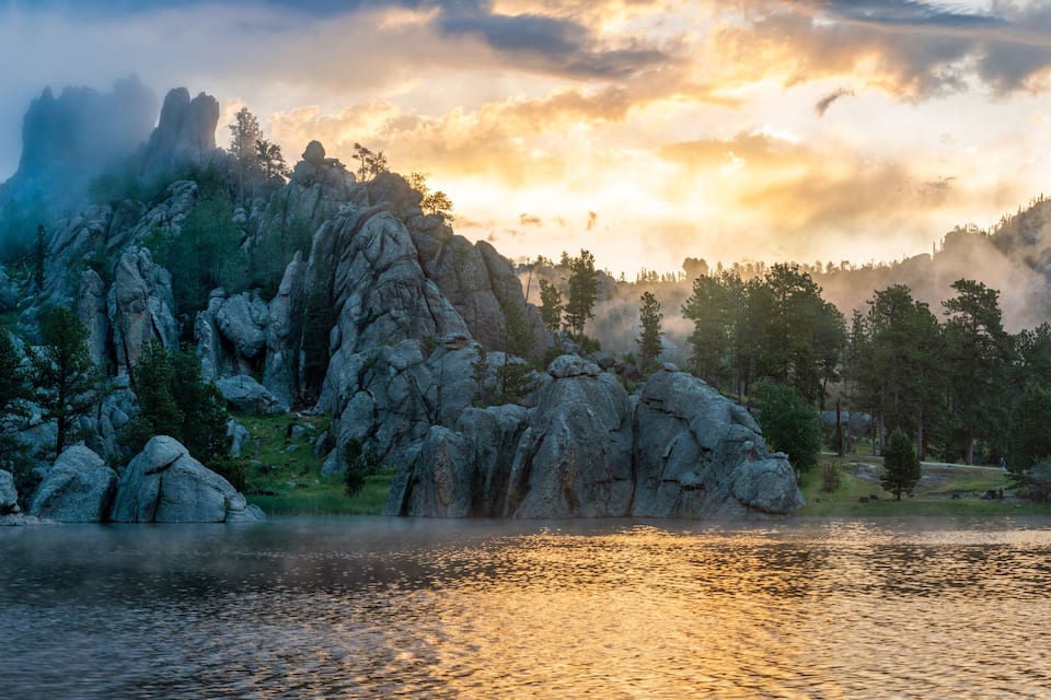 A mountainous landscape with a lake reflecting the sky, surrounded by dense forests and rocky outcrops.