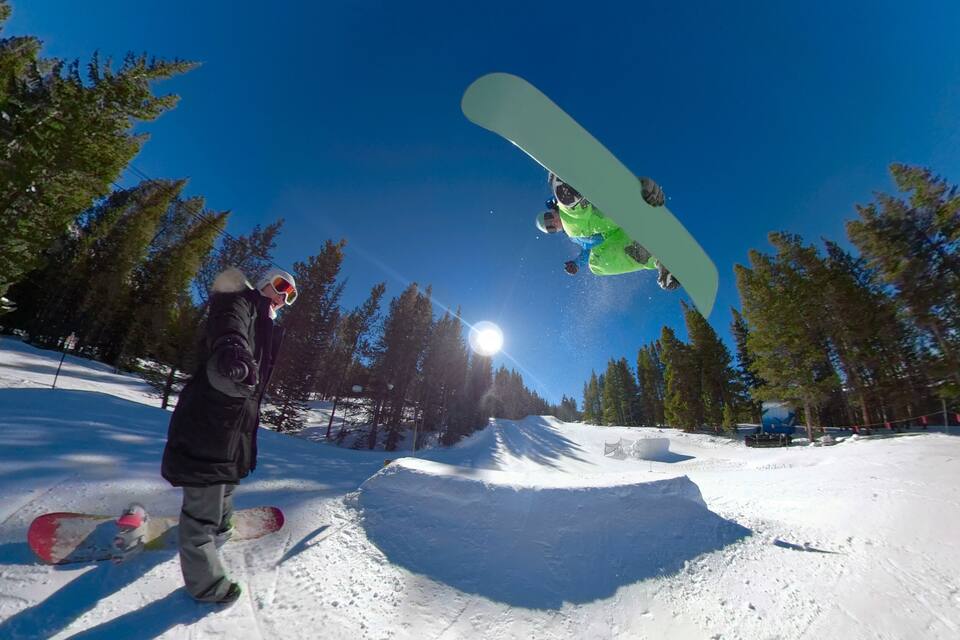 Un snowboarder en el aire realizando un truco en una ladera nevada, con árboles al fondo.