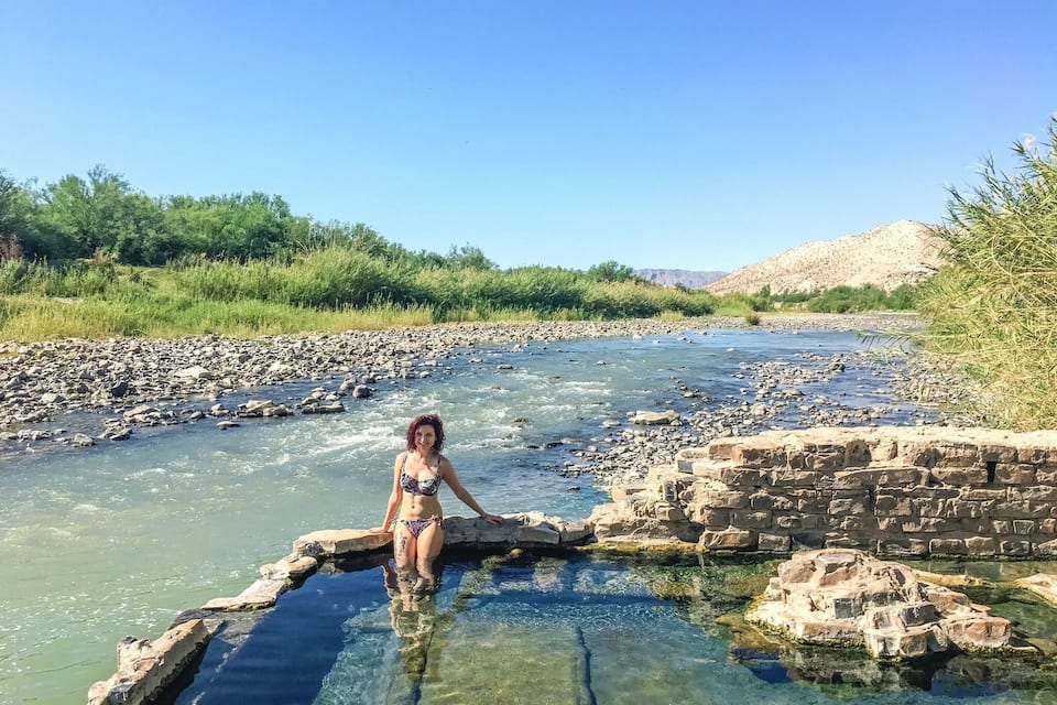 Una mujer con traje de baño sentada en un río cristalino con orillas rocosas y follaje al fondo.