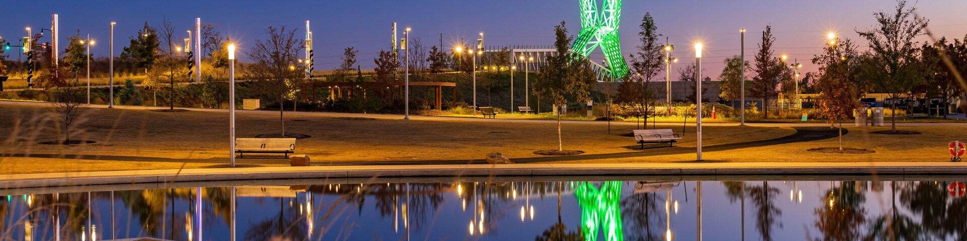 Cena noturna com uma escultura iluminada em verde ao longe, um corpo d'água calmo refletindo luzes e postes de rua ao longo da área.