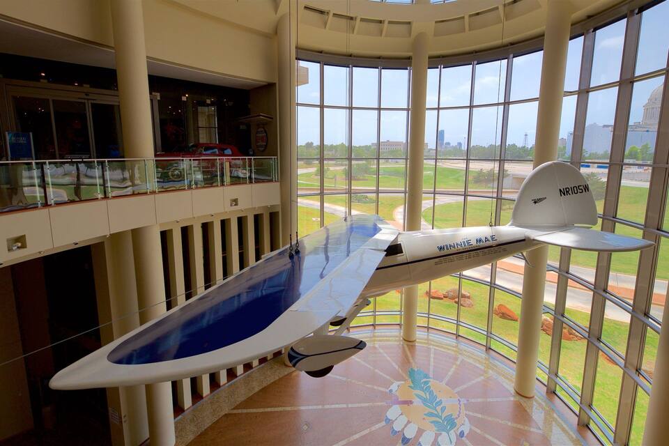 A model airplane named 'Winnie Mae' suspended in a building with large windows overlooking a cityscape.
