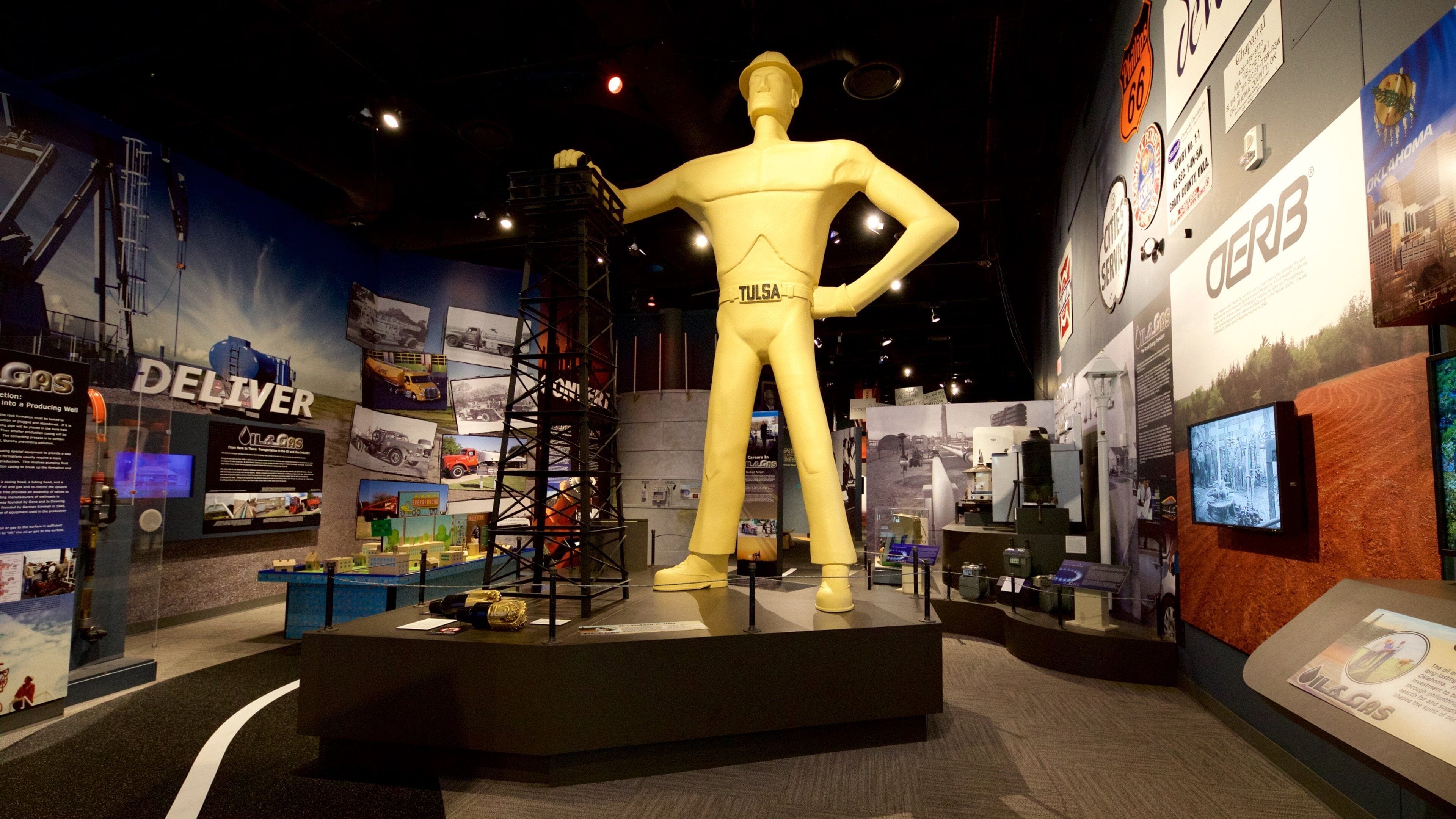 Una estatua grande y amarilla de un hombre con traje, con la palabra 'TULSA' en la espalda, rodeada de paneles informativos y exhibiciones.