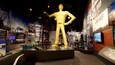 Una estatua grande y amarilla de un hombre con traje, con la palabra 'TULSA' en la espalda, rodeada de paneles informativos y exhibiciones.
