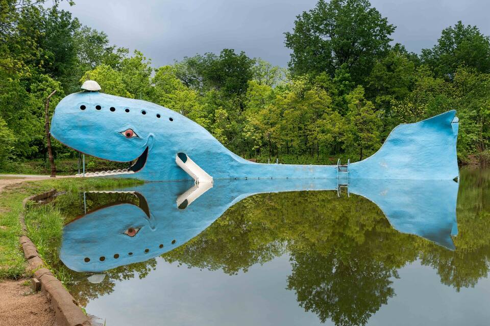A large blue sculpture of a whale with a smiling face, reflecting in a body of water, surrounded by trees.