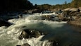 Ein Fluss mit schnell fließendem Wasser über Felsen, umgeben von bewaldeten Hügeln.