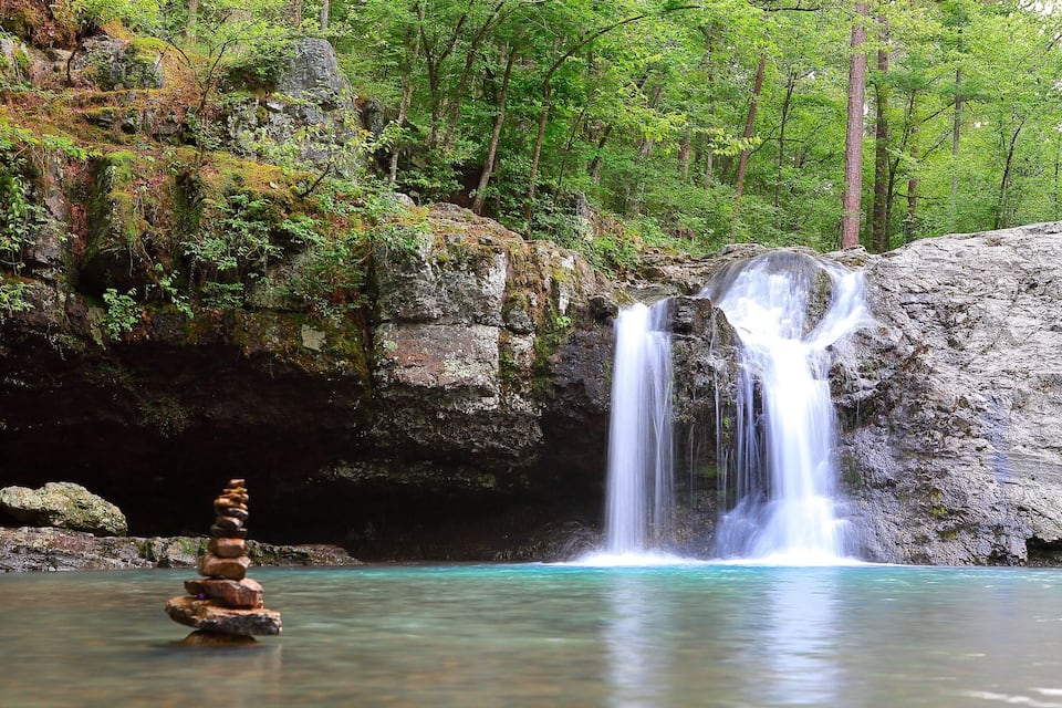 A waterfall cascading into a clear pool surrounded by forested rocks and moss-covered boulders.
