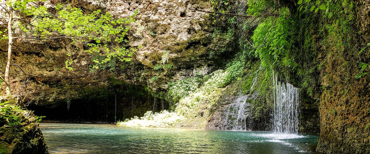 A serene waterfall cascading into a clear pool surrounded by lush greenery and rocky cliffs.
