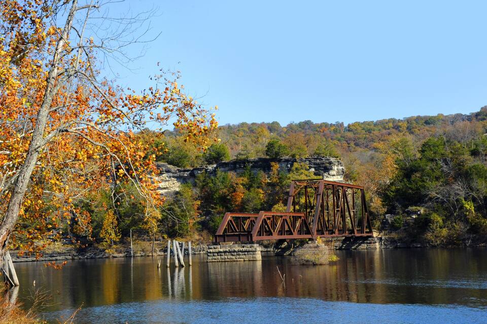 A rusted metal bridge over a calm river, surrounded by autumn foliage and rocky cliffs.