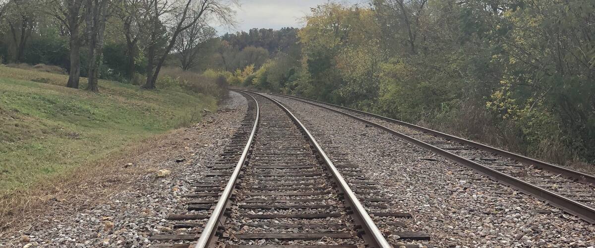 Railway tracks curving through a wooded area with a cloudy sky.