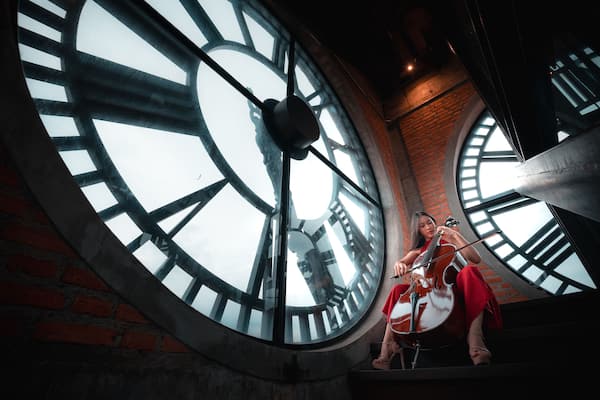 A woman sits in front of a clock with a cello. The clock is large and has a very old-fashioned look. The woman is dressed in red and is playing the cello. The image has a vintage