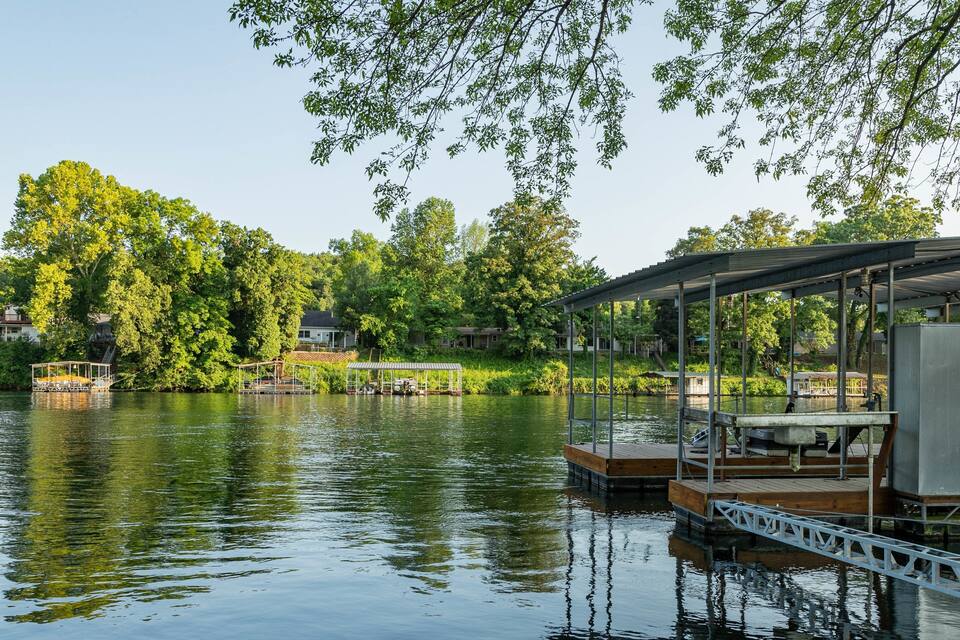 A calm lake with docked boats, a covered seating area, and houses in the background.