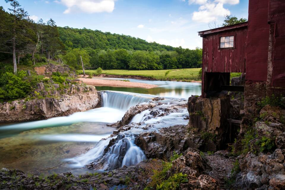 Un cascada, un riu i un edifici vermell en una zona boscosa.