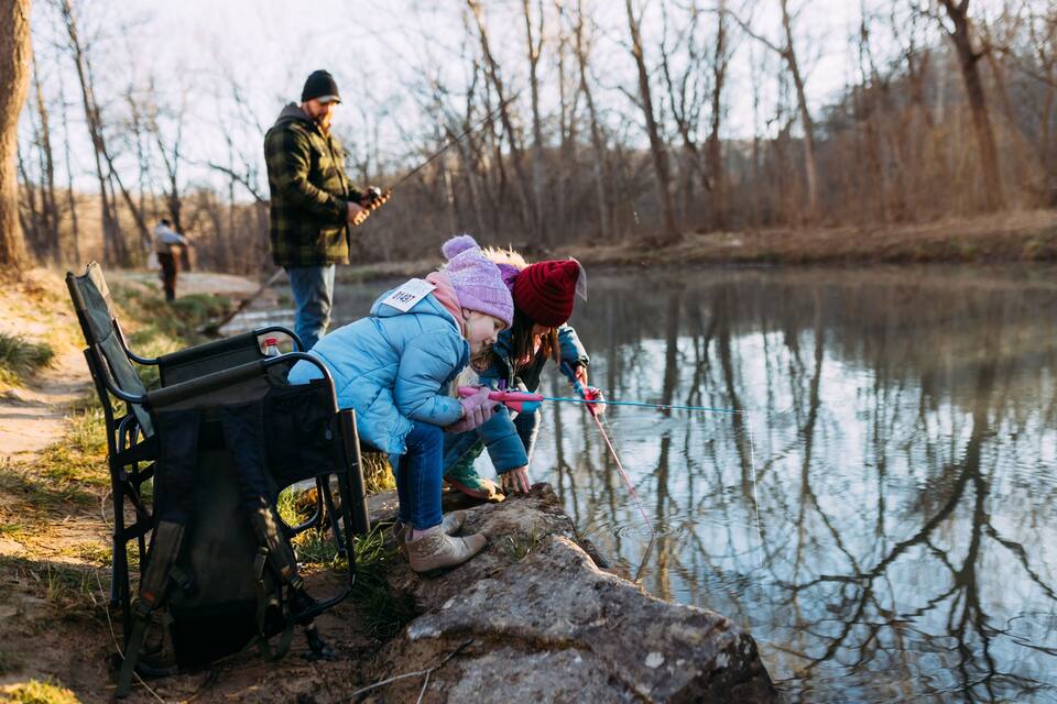 A group of people fishing by a calm river with trees in the background.