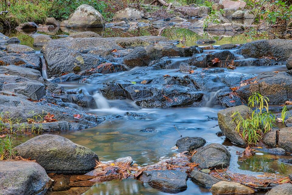 A rocky stream with clear water, surrounded by large boulders and autumn leaves.