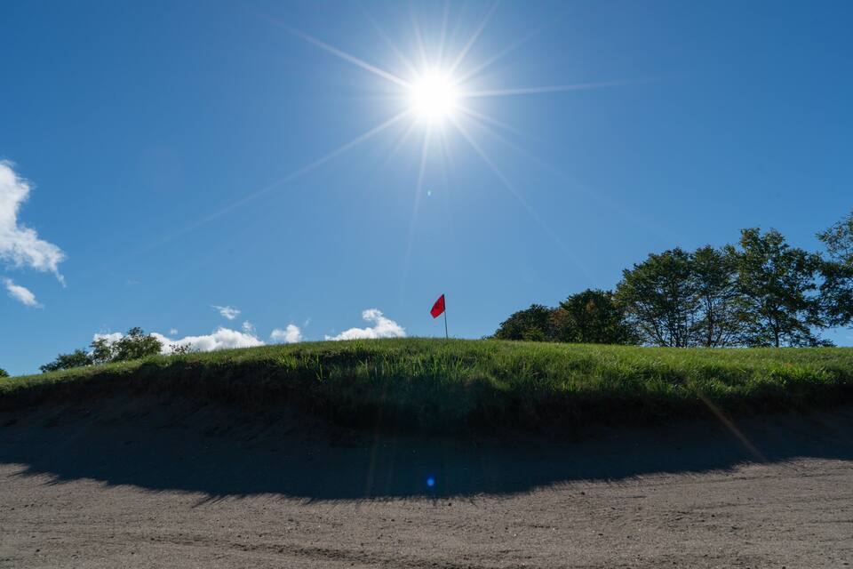 A clear blue sky with the sun shining brightly, a red flag on a grassy hill, and a dirt path leading towards it.