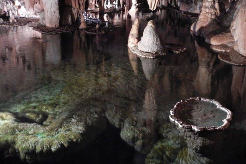 A cave with stalactites, stalagmites, and a reflective water surface.