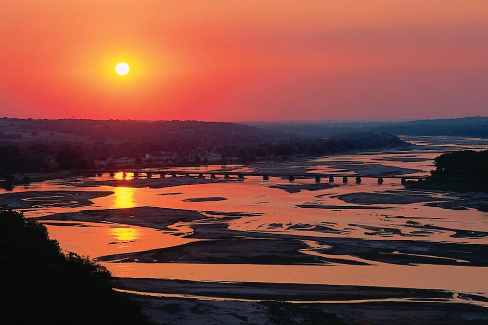 A sunset over a river with a bridge and surrounding hills.