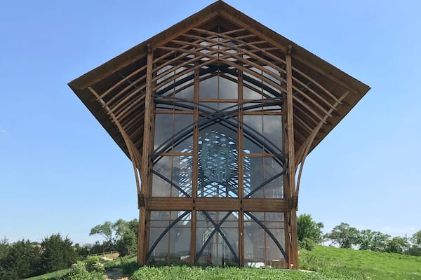 A modern glass structure with a wooden frame, set in a grassy field with trees in the background.