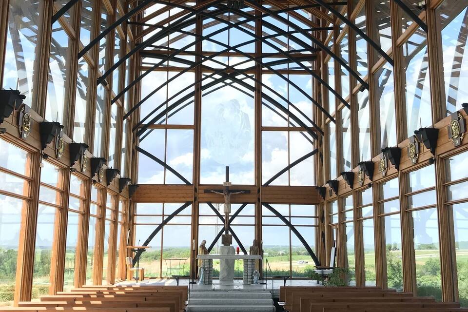 Ein Kircheninneres mit Holzbänken, einem Kruzifix und Blick auf die Landschaft durch große Fenster.